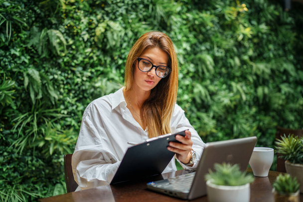 One female young adult with long hair and glasses caucasian sitting in restaurant or hotel working and prepare for business meeting online using digital tablet and paper documents copy space - Photo, Image