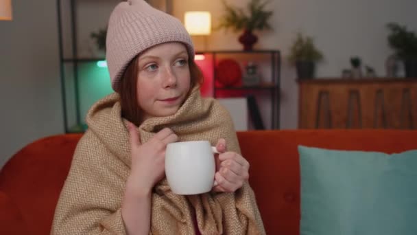 Sick child girl wear hat wrapped in plaid sit alone shivering from cold on sofa drinking hot tea in unheated apartment without heating due debt. Unhealthy children feeling discomfort try to warming up - Filmati, video