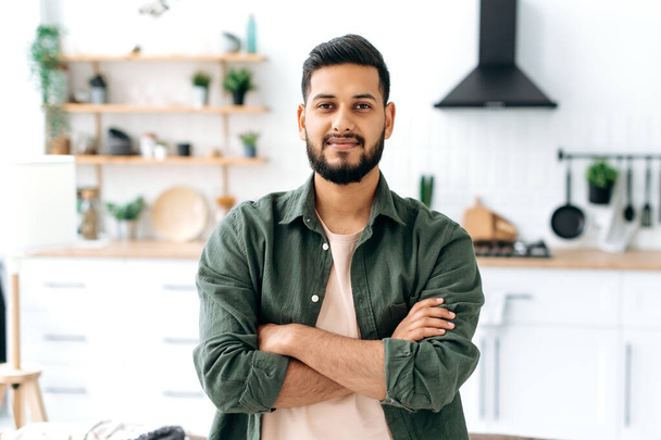 Красивий впевнений привабливий сучасний індійський або арабський хлопець, в повсякденному стильному одязі, що стоїть вдома у вітальні на фоні кухні, з схрещеними руками, дивиться на камеру, посміхається
 - Фото, зображення