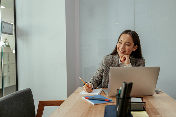 Lächelnde asiatische Geschäftsfrau, die im Büro arbeitet, benutzt Computer und macht sich Notizen. Unklarer Hintergrund - Foto, Bild