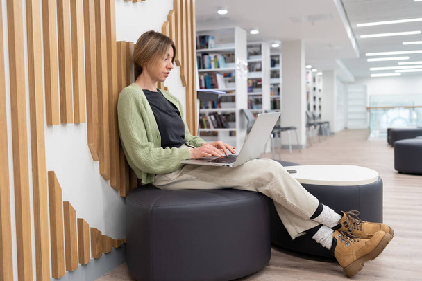 Middle-aged woman freelancer wearing casual clothes working on laptop computer in library, mature female student typing on keyboard while studying online in reading room, work from anywhere. - Foto, Bild