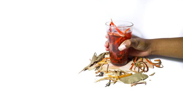 La mano de un hombre asiático sostiene un vaso de bebida herbal, con algunas especias a su alrededor. bebida a base de hierbas aislada sobre un fondo blanco - Foto, imagen