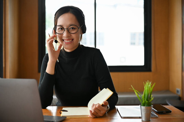 Όμορφη και χαρούμενη νεαρή φοιτήτρια με ακουστικά και γυαλιά μελετώντας ένα online μάθημα μέσω φορητού υπολογιστή. - Φωτογραφία, εικόνα