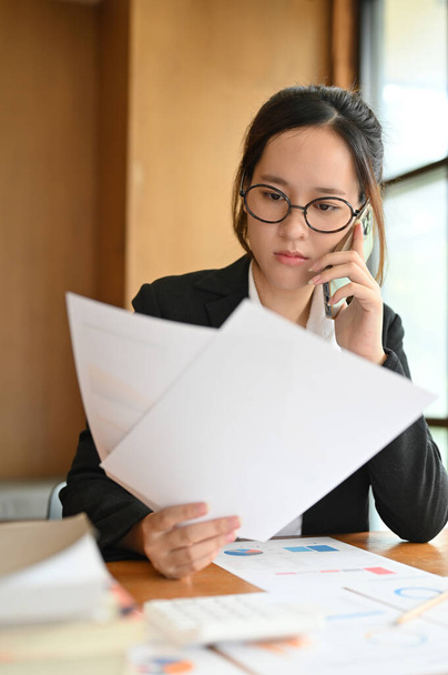 Professional young Asian female accountant or financial worker focused reading something on paperwork while on the phone call with her client. - Foto, imagen