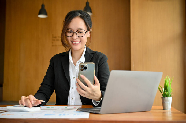 Όμορφη νεαρή Ασιάτισσα λογίστρια με γυαλιά κάθεται στο γραφείο της και χρησιμοποιεί ένα smartphone για να ψάξει κάποιες πληροφορίες στο διαδίκτυο. - Φωτογραφία, εικόνα