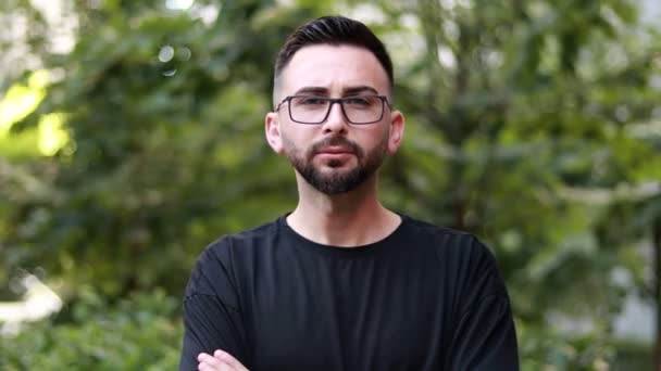 Portrait of young handsome man with beard and glasses. Young man in black shirt smiling looking at camera. Outdoor nature background - Video