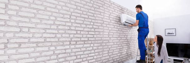 Young Woman Looking At Male Technician Fixing Air Conditioner On Brick Wall - Fotoğraf, Görsel