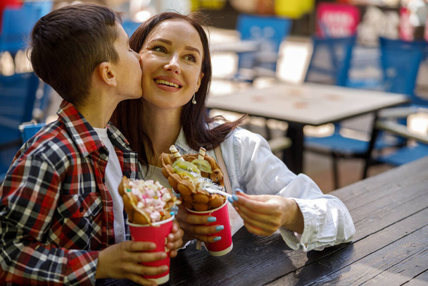 Cute little son kissing pretty mother and eating sweet waffles on weekend summer day. Fast food. - Foto, Imagem