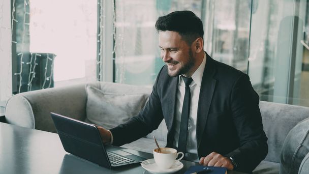 Cheery bearded Caucasian businessman in formal clothes chatting to his family through laptop and waving his hand happily in a classy cafe. He gesticulates optimistically and cheerfully. - 写真・画像
