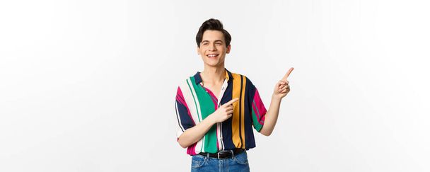 Безтурботний хлопець у стильній сорочці, спрямованій пальцями у верхньому правому куті, показує рекламний банер або логотип, посміхається і розмовляє з кимось, стоїть над білим тлом
. - Фото, зображення