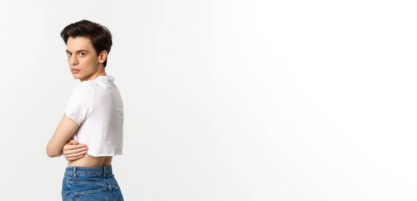 Lgbtq and pride concept. Profile view of beautiful gay man in crop top turn head at camera, staring with sensual gaze, standing over white background. - Photo, Image