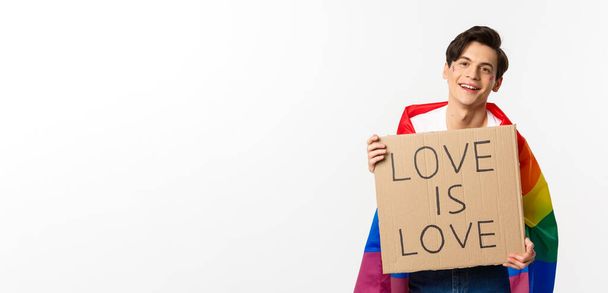 sorridente gay uomo attivista holding segno amore è amore per lgbt orgoglio parata indossare arcobaleno bandiera in piedi su bianco sfondo. - Foto, immagini
