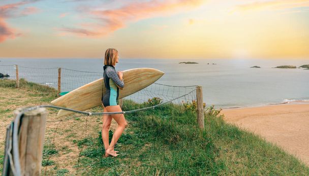 Felismerhetetlen fiatal szörfös nő szörfös öltönyben és szörfdeszkával, aki a partról nézi a strandot - Fotó, kép