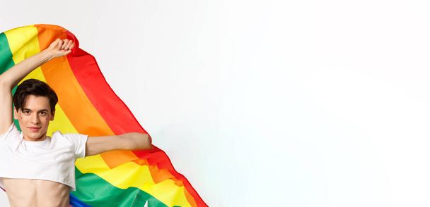 Vertikale Ansicht einer glücklichen queeren Person in bauchfreiem Top und Jeans, die eine hochgezogene Regenbogenfahne schwenkt, um den lgbtq-Feiertag zu feiern, auf weißem Hintergrund stehend. - Foto, Bild
