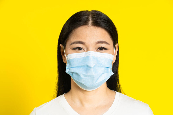 Happy, safe of pandemic mature asian woman wearing medical face mask coronavirus or monkeypox prevention. Charming middle age woman in white t-shirt and medical mask on yellow background.  - Foto, Imagem