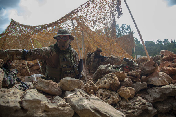 A team of airsoft players hid behind a stone protective structure covered with a camouflage net. The squad leader gives instructions with his hand. Paramilitary Detachment - 写真・画像