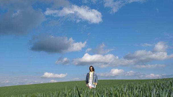 Krásná dívka s dlouhými vlasy oblečená v džínové bundě kráčí sama po zeleném poli. Šťastná, svobodná mladá dívka v zelené pšenici. - Fotografie, Obrázek