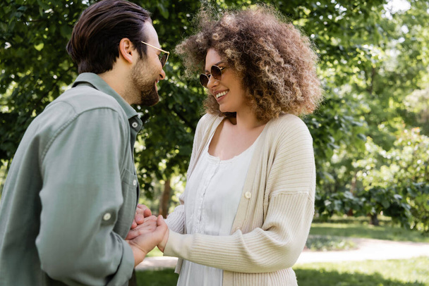 curly woman holding hands with happy man in stylish sunglasses in park - Foto, afbeelding