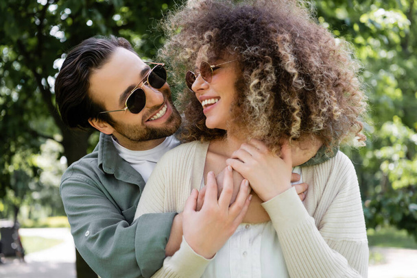 happy man in stylish sunglasses hugging pleased girlfriend in park - Photo, image
