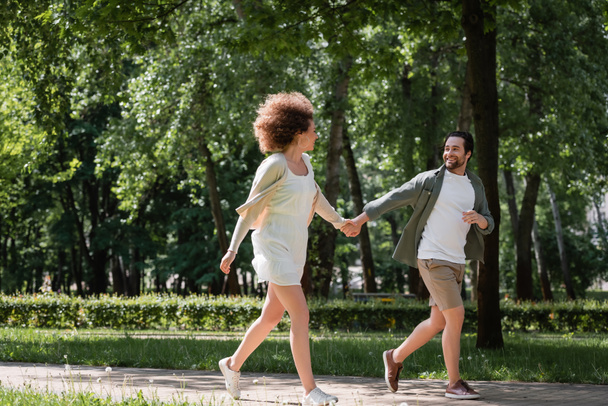 teljes hossza pozitív fiatal pár fogja egymás kezét, miközben fut együtt a nyári parkban  - Fotó, kép