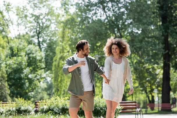joyful young couple holding hands while walking together in summer park  - 写真・画像