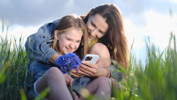 公園内で自然を満喫している美しい若い母親と娘たちは、スマートフォンを使って笑顔で楽しんでいます。陽気な家族 - 写真・画像