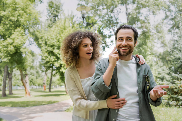 curly woman hugging near happy boyfriend talking on smartphone in green park - Photo, Image