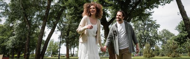 весёлый мужчина и довольная кудрявая женщина в платье держась за руки в летнем парке, баннер - Фото, изображение