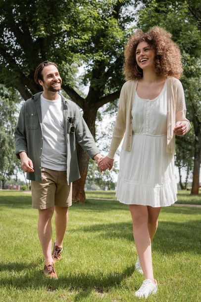 joyful man and pleased curly woman in dress holding hands in summer park  - Photo, image