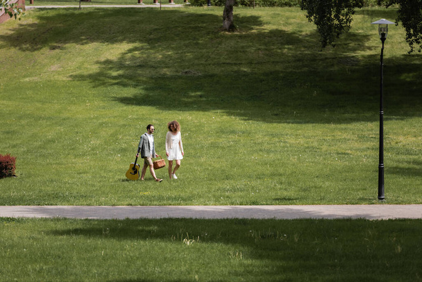 full length of man in summer clothes carrying guitar and picnic wicker basket near happy girlfriend in green park  - Fotografie, Obrázek