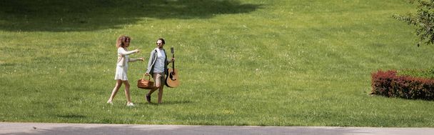 full length of man carrying guitar and wicker basket near happy girlfriend gesturing in green park, banner - Zdjęcie, obraz