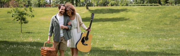 happy curly woman holding acoustic guitar near man with wicker basket and walking in green park, banner - Foto, imagen