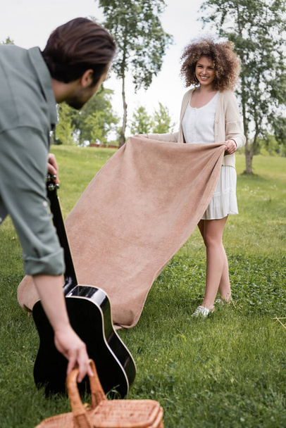 cheerful curly woman in summer clothes holding blanket near boyfriend with acoustic guitar in park - Foto, imagen