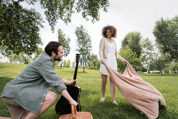pleased curly woman in summer clothes holding blanket near boyfriend in park - Photo, Image
