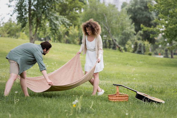 cheerful young couple in summer clothes putting picnic blanket on green grass in park - Foto, immagini