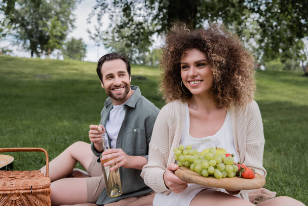 cheerful woman holding cutting board with fresh fruits near happy boyfriend opening bottle of wine  - Foto, Bild