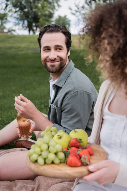happy man opening bottle of wine near curly girlfriend sitting with fruits during picnic - 写真・画像