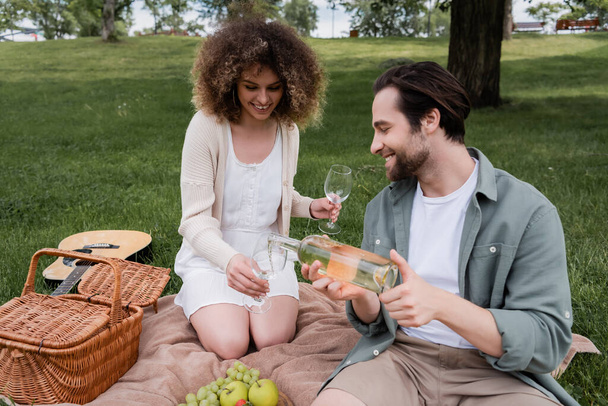 cheerful man pouring wine in glass near curly woman during summer picnic - Foto, Imagem