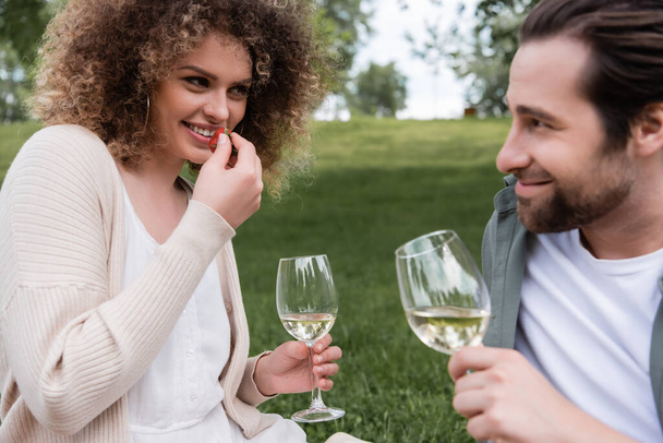 happy curly woman with glass of wine eating fresh strawberry near boyfriend  - Fotó, kép