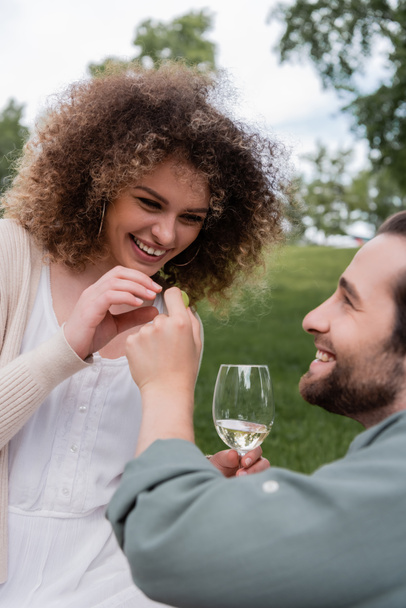 happy man with glass of wine holding fresh grape near cheerful girlfriend during picnic - Fotó, kép