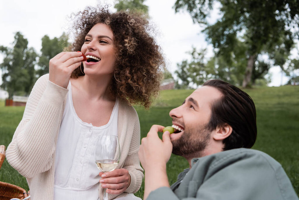 casal feliz comer frutas frescas perto de copo de vinho durante piquenique - Foto, Imagem
