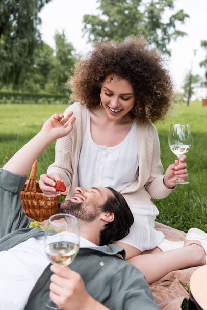 cheerful curly woman holding glass of wine and feeding boyfriend with fresh strawberry during picnic - Valokuva, kuva