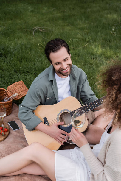 high angle view of romantic man playing acoustic guitar near curly woman with glass of wine during picnic  - Zdjęcie, obraz
