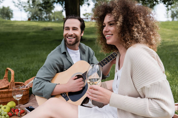 Χαρούμενος άντρας παίζει ακουστική κιθάρα κοντά ευτυχισμένη γυναίκα με ένα ποτήρι κρασί κατά τη διάρκεια του πικ-νικ  - Φωτογραφία, εικόνα