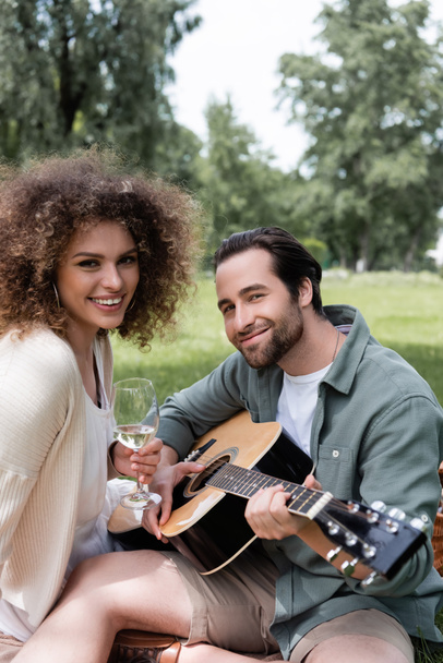 hombre romántico tocando la guitarra acústica cerca de la mujer sonriente con copa de vino durante el picnic  - Foto, imagen