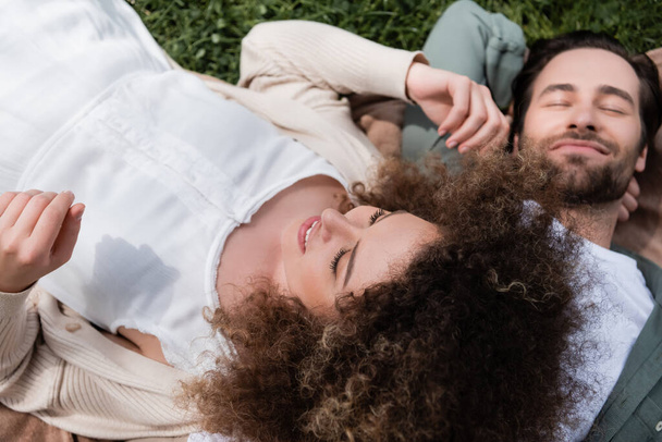 high angle view of curly woman lying on pleased boyfriend during picnic in summer park - Foto, afbeelding