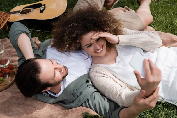 vista superior de la mujer feliz tomando selfie en el teléfono inteligente mientras está acostado con su novio durante el picnic - Foto, Imagen