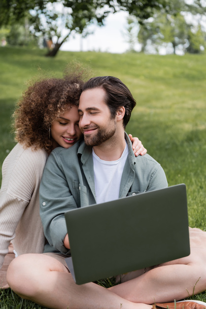 curly woman smiling while hugging boyfriend with laptop working remotely in park - Photo, image
