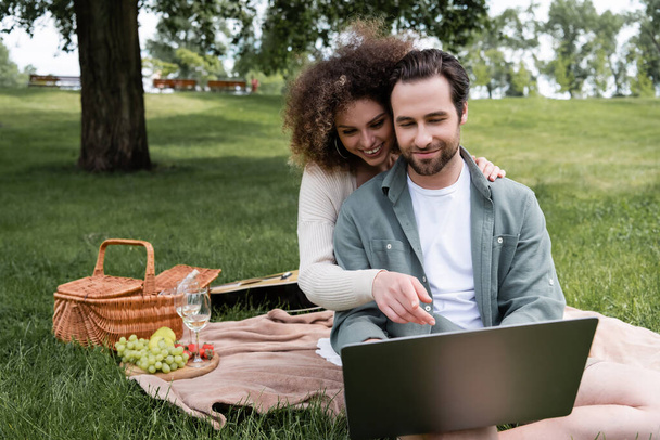 curly woman pointing at laptop near boyfriend during picnic in park - Foto, Bild