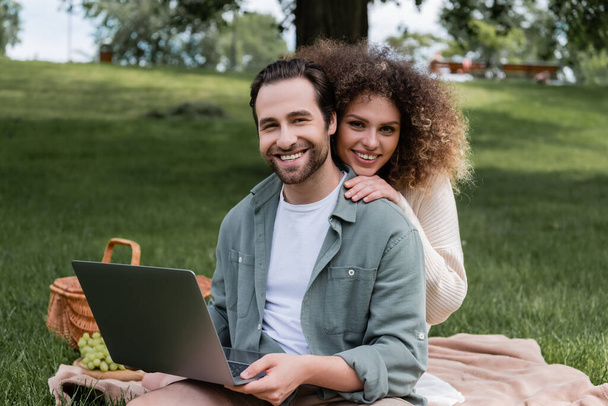 веселый мужчина держит ноутбук и улыбается с кудрявой женщиной во время пикника - Фото, изображение